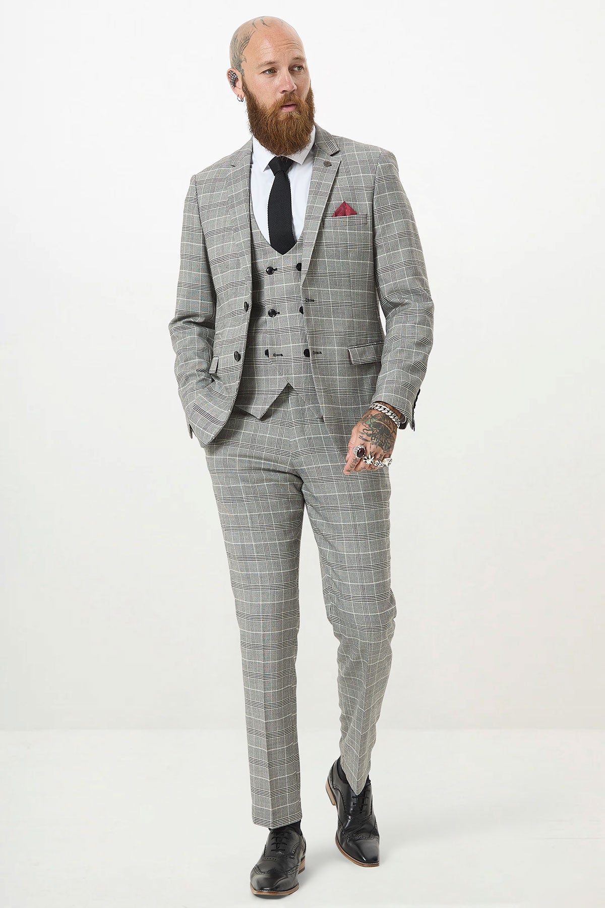 A bearded, tattooed man wears the Marc Darcy ROSS Grey Check Three Piece Suit with a double-breasted waistcoat, white shirt, black tie, black shoes, and a red pocket square as he strides confidently against a plain white background.