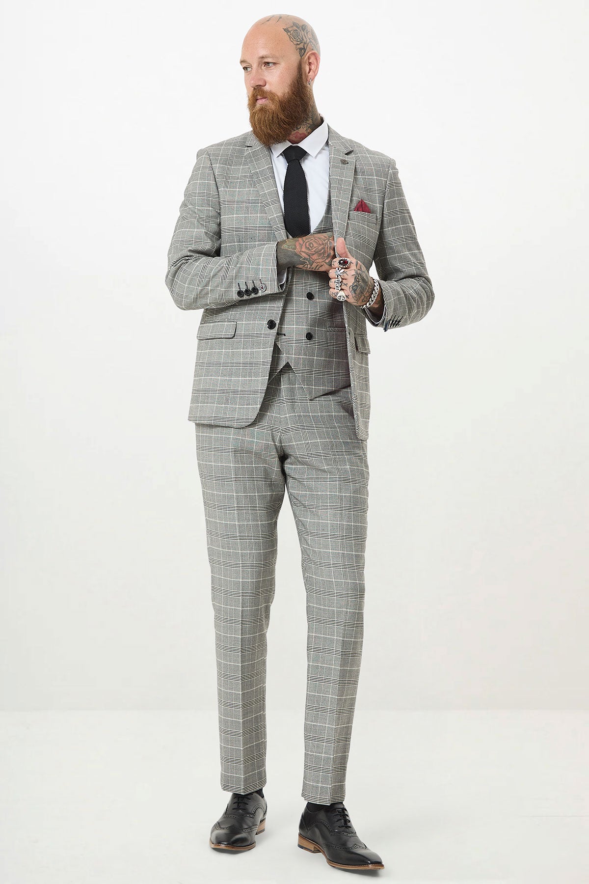 A bearded man with tattoos wears the Marc Darcy ROSS Grey Check Three Piece Suit with a white shirt, black tie, and black dress shoes. He stands against a plain white background, adjusting his double-breasted waistcoat.