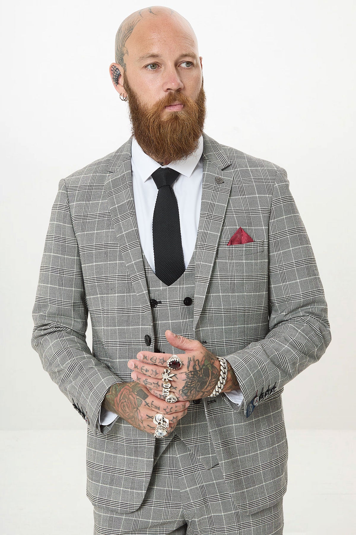 A bearded, tattooed man stands against a white background wearing the Marc Darcy ROSS Grey Check Three Piece Suit with a white shirt, black tie, and red pocket square, hands together.