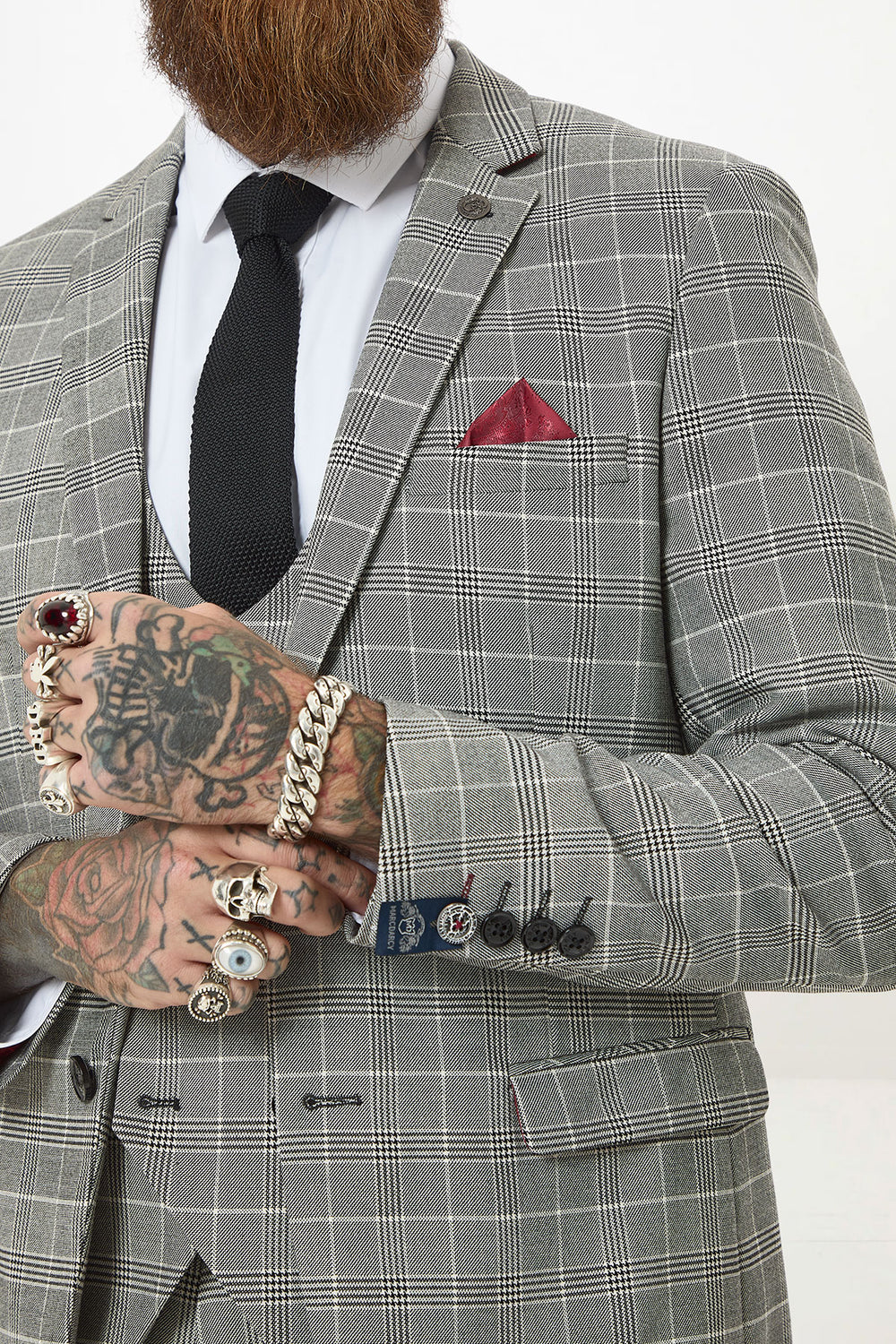 A man wears the Marc Darcy ROSS Grey Check Three Piece Suit with a black tie and red pocket square. His tattooed, ringed hands are clasped, and he has a brown beard. The photo is cropped at his chin.