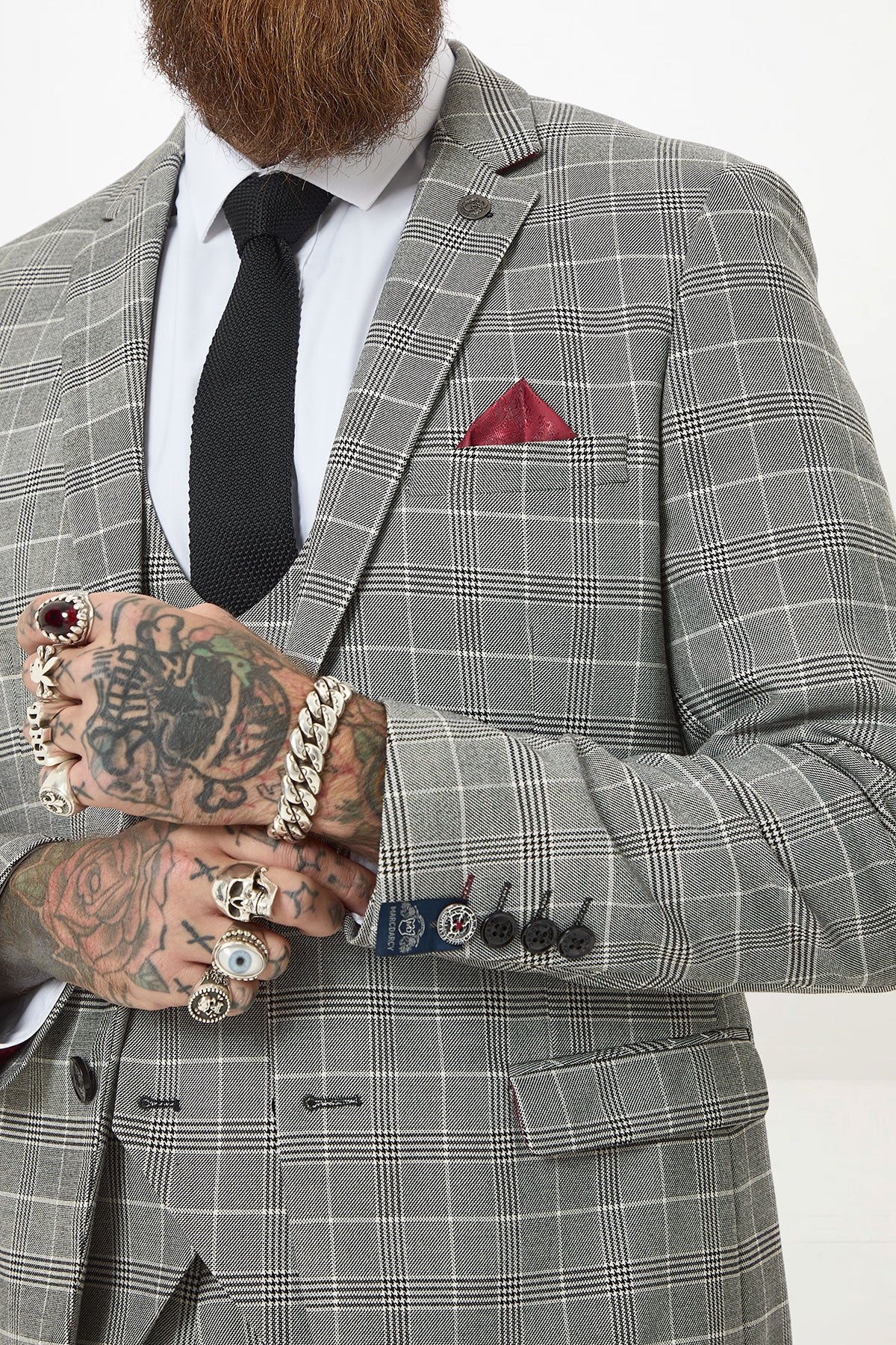 Wearing the Marc Darcy ROSS Grey Check Three Piece Suit, a person pairs it with a white shirt, black tie, and red pocket square. Their tattooed, ring-adorned hands are clasped, blazer sleeves slightly raised. The face is partly out of frame.