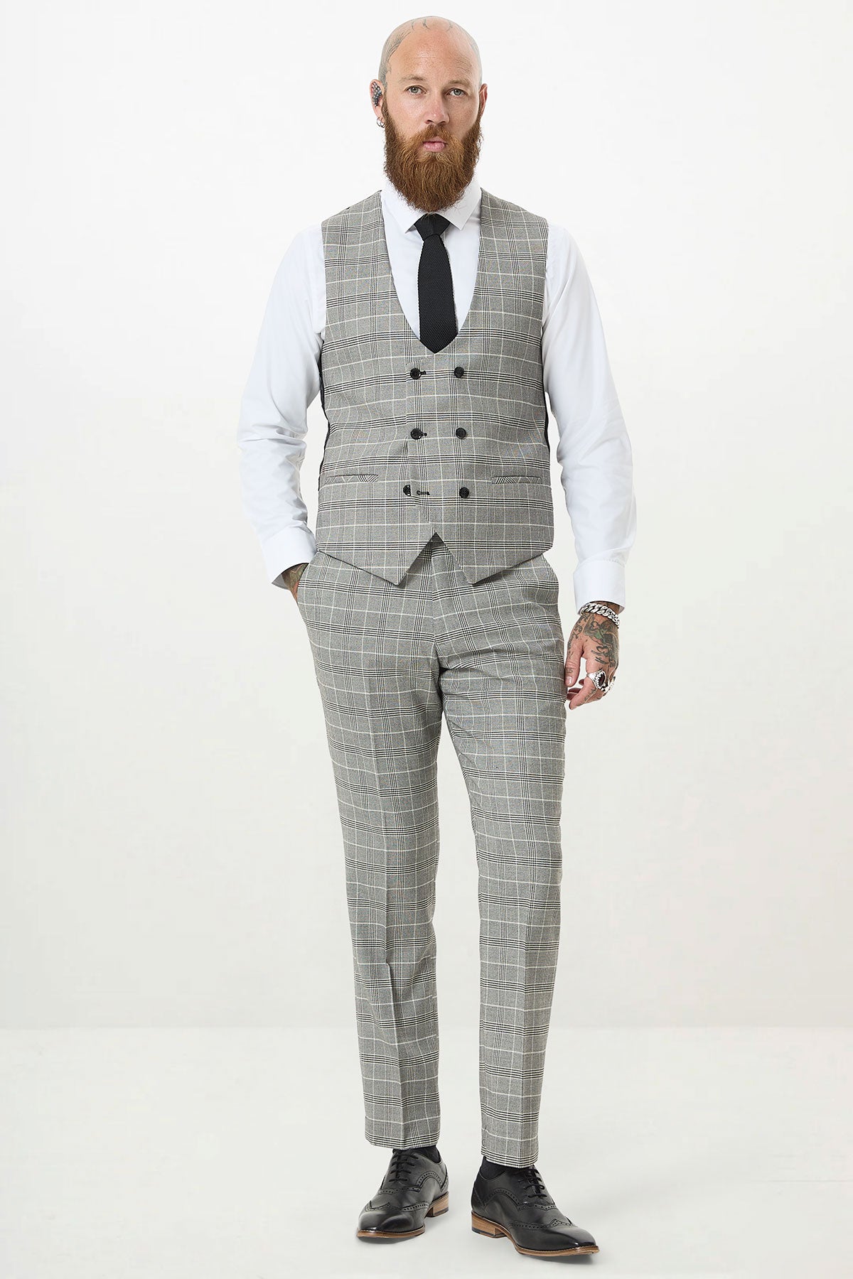 A bearded, tattooed man stands facing forward in a Marc Darcy ROSS Grey Check Three Piece Suit with a white shirt, black tie, and black dress shoes against a plain white background.