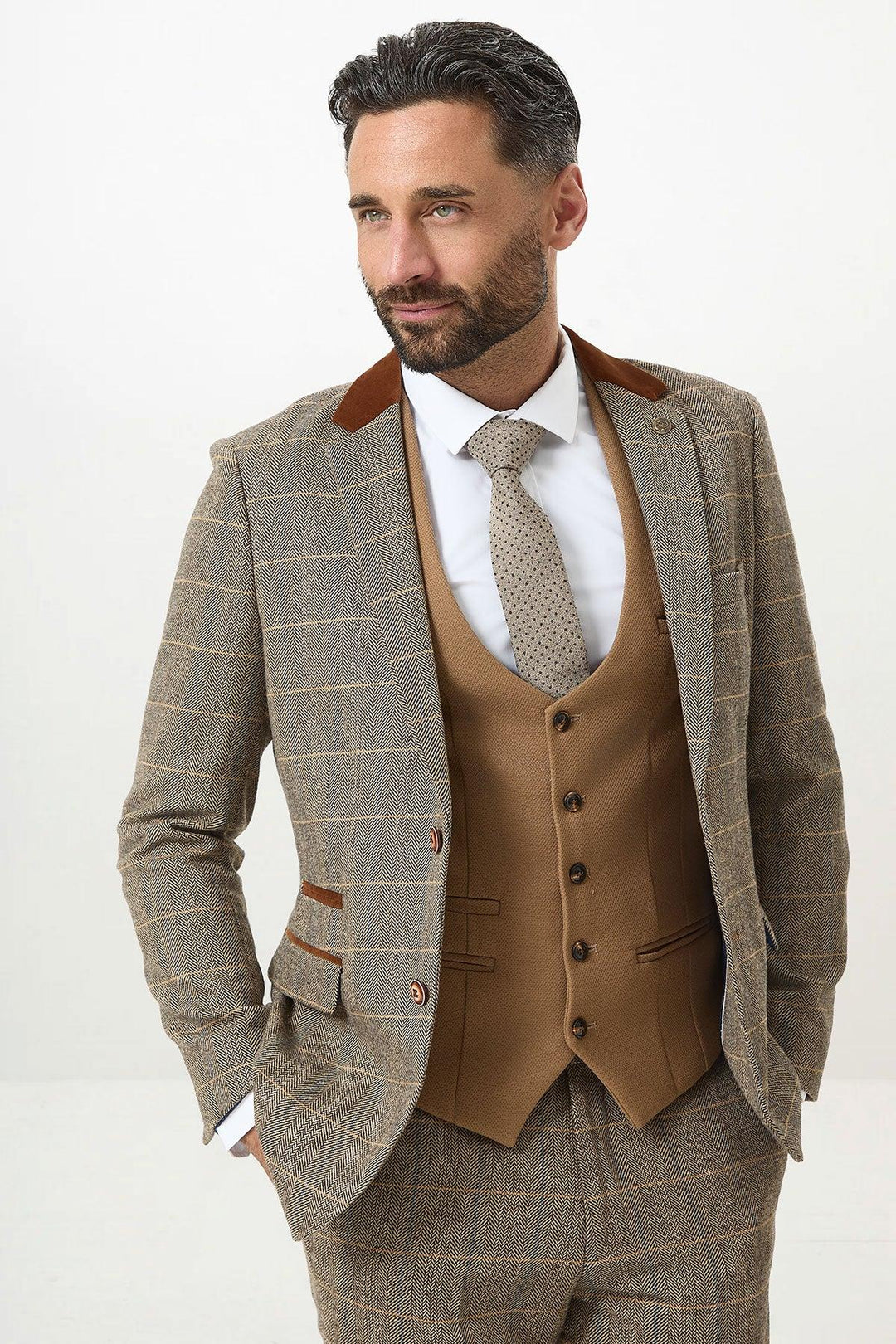A man with dark hair and a beard poses with hands in his pockets, wearing the marcdarcy DX7 - Tan Tweed Check Suit With Kelvin Oak Waistcoat, styled with a white shirt and patterned tie.