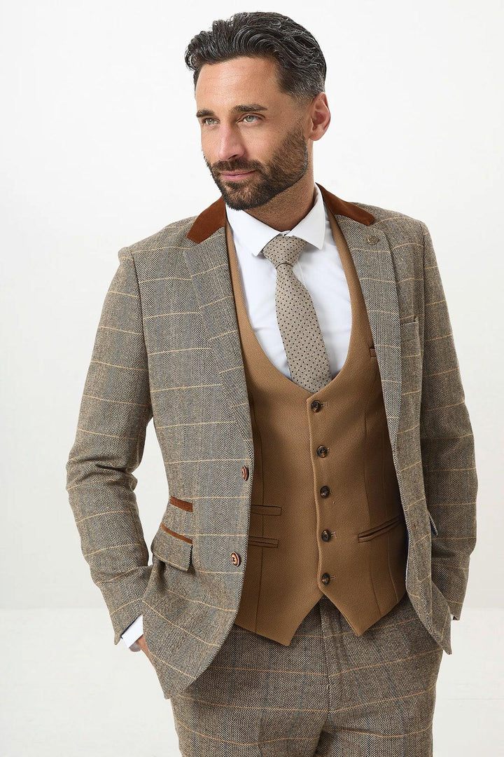 A man with dark hair and a beard poses with hands in his pockets, wearing the marcdarcy DX7 - Tan Tweed Check Suit With Kelvin Oak Waistcoat, styled with a white shirt and patterned tie.
