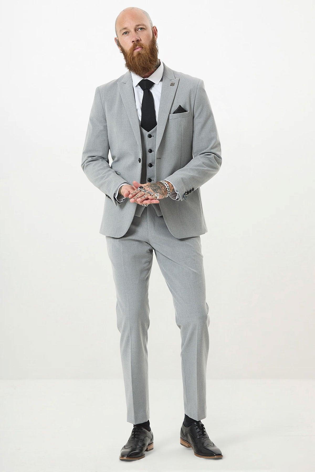 A bearded man with hand tattoos wears the Marc Darcy BROMLEY Silver Grey Check Three Piece Suit, paired with a white shirt, black tie, and black shoes, standing against a plain white background.