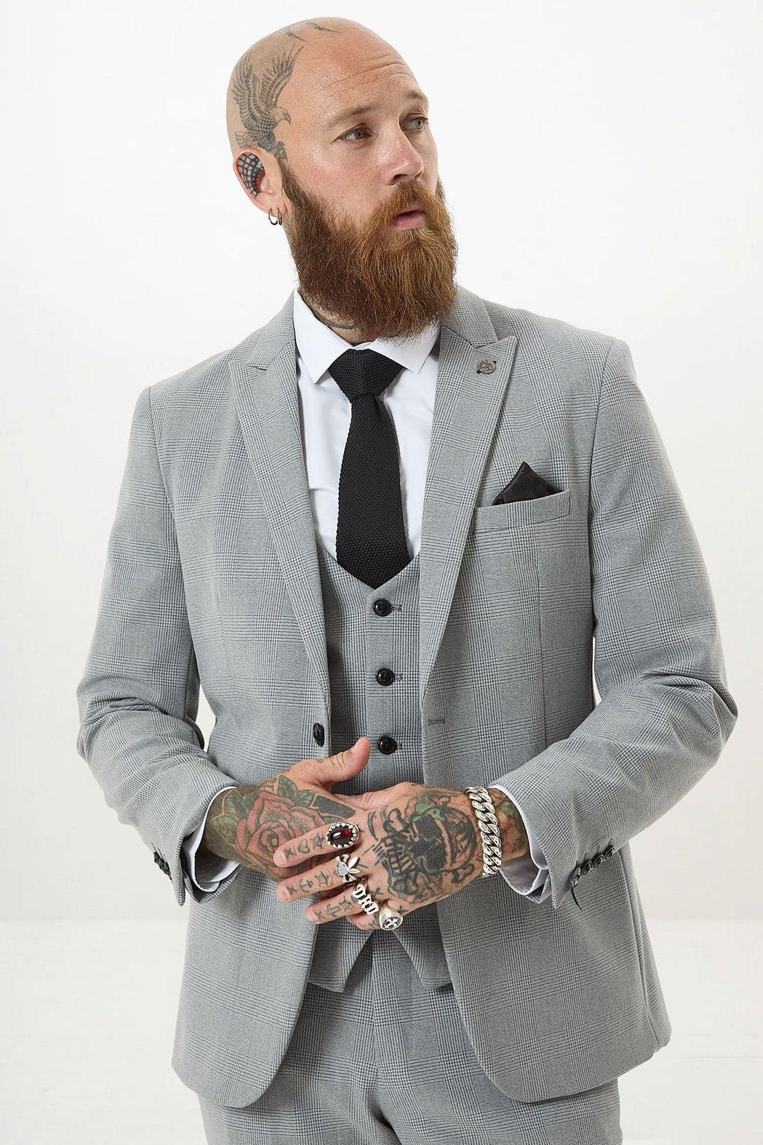 A bearded man with tattoos and piercings wears the Marc Darcy BROMLEY Silver Grey Check Blazer, paired with a white shirt and black tie. He looks to the side, adjusting his suit against a plain white background.