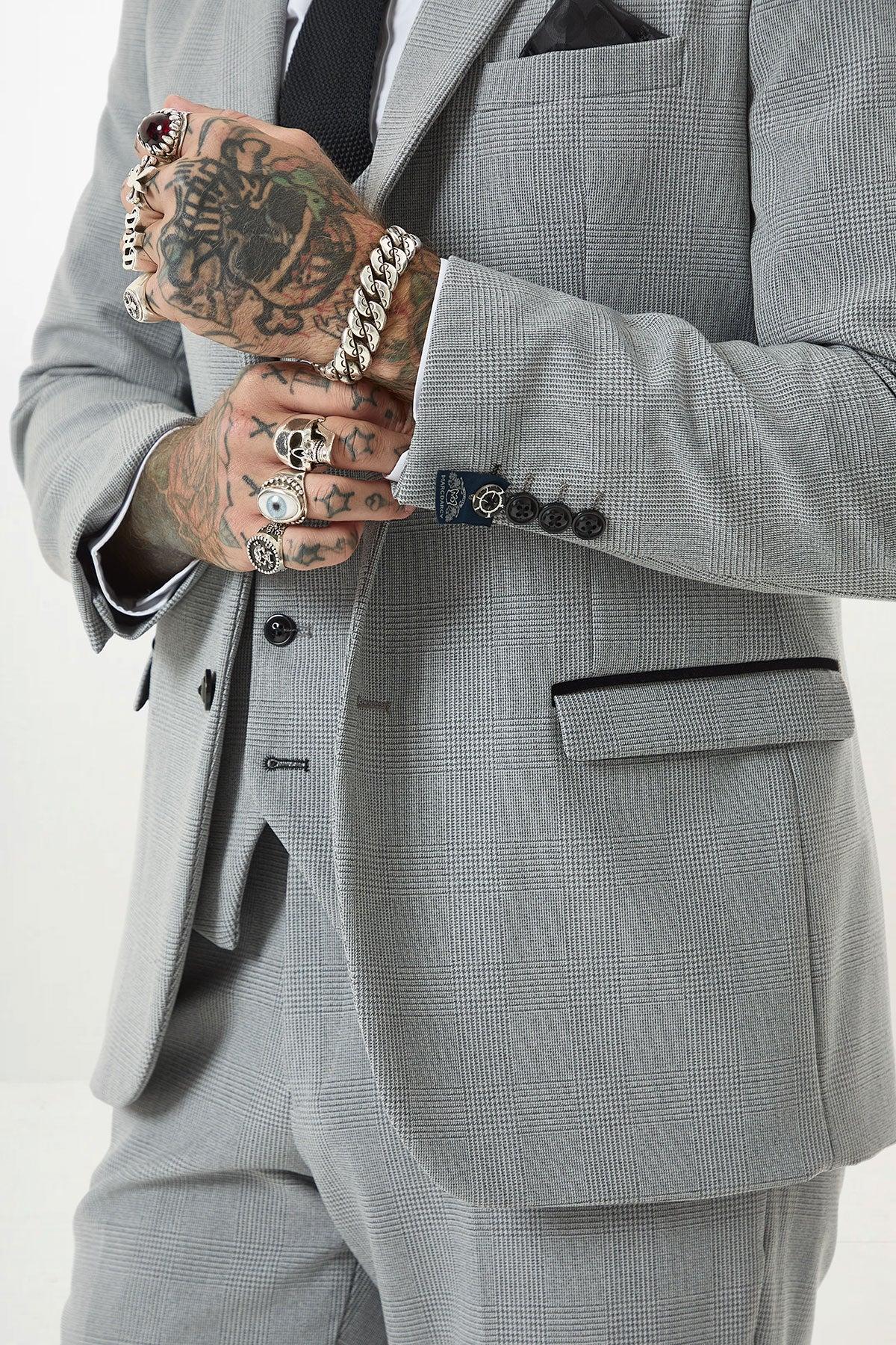 A person wears the Marc Darcy BROMLEY Silver Grey Check Three Piece Suit with black-trimmed pockets and buttons, paired with a black tie. Their heavily tattooed hands display multiple rings and a thick silver bracelet.