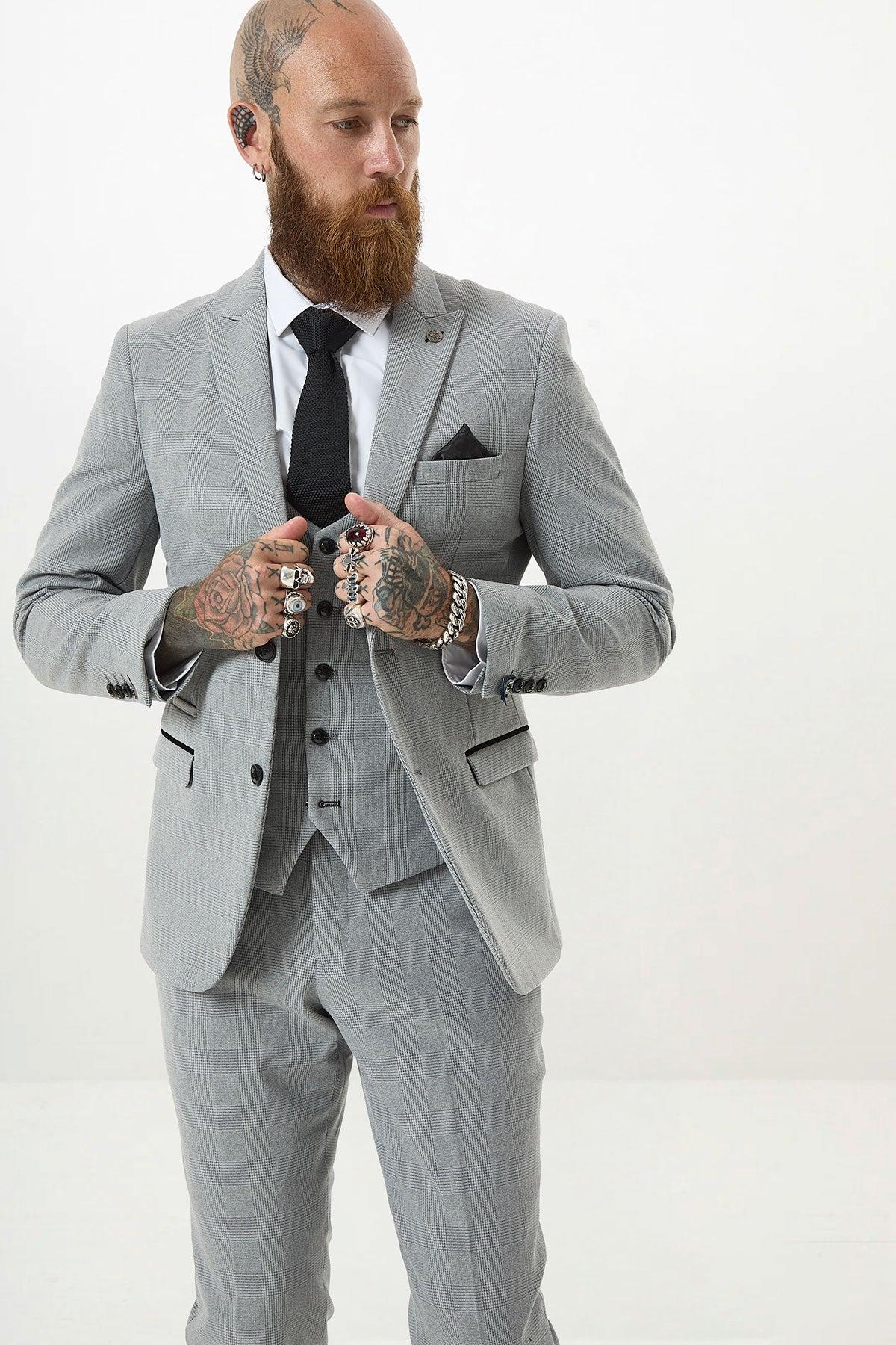 A bearded, tattooed man wears the Marc Darcy BROMLEY - Silver Grey Check Blazer, styled slim fit with a black tie and pocket square. He adjusts his jacket while looking to the side against a plain white background.