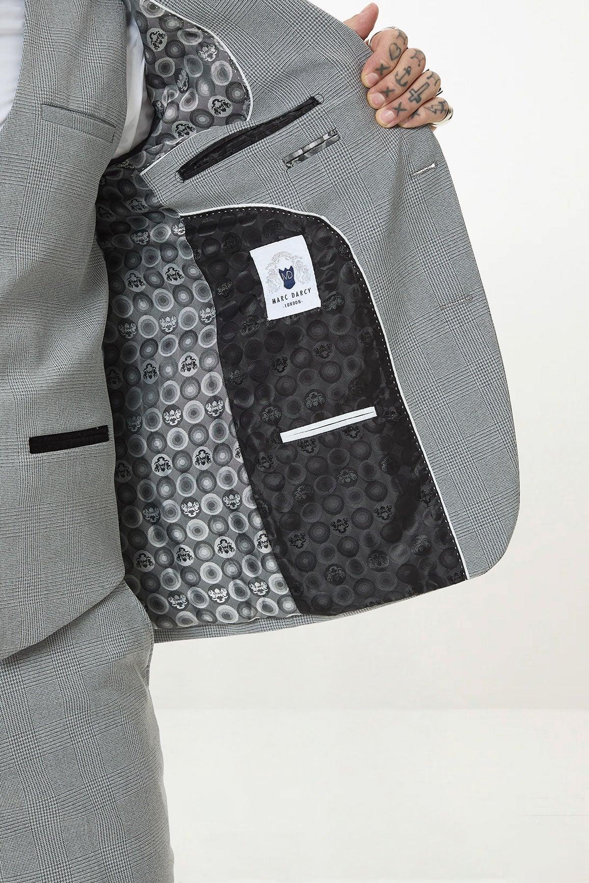 A person holds open a slim fit BROMLEY Silver Grey Check Three Piece Suit by Marc Darcy, showing a black inner lining with a skull pattern and a white "Marc Darcy" label. Tattoos can be seen on the person's fingers.
