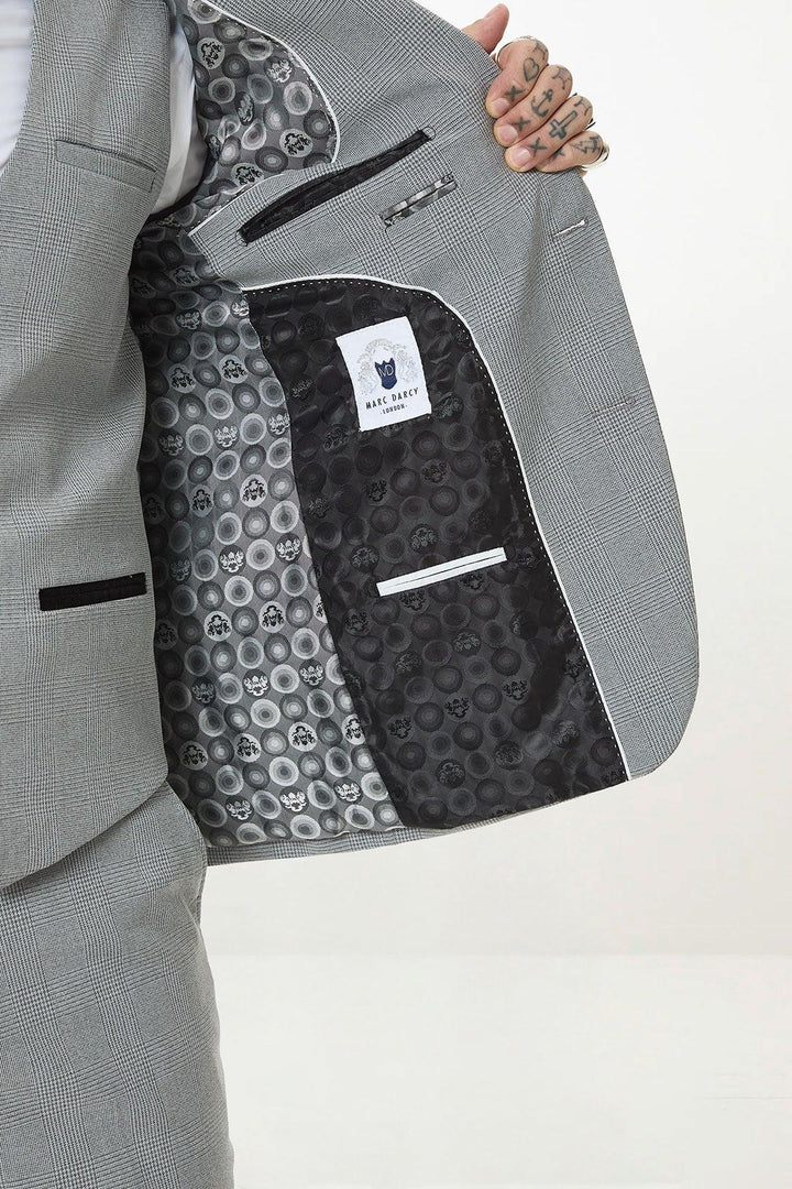 A person holds open the BROMLEY - Silver Grey Check Blazer by Marc Darcy, showing a black skull-patterned lining and white Marc Darcy label. The Prince of Wales check design adds style, with tattoos visible on their fingers.