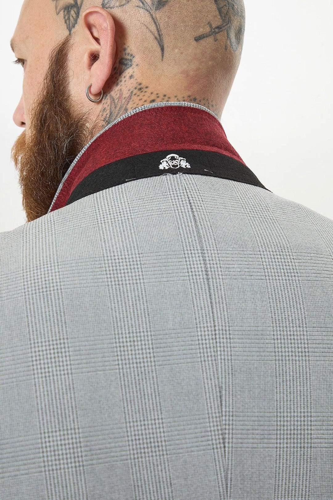 A close-up of a bearded man with tattoos on his shaved head, wearing the Marc Darcy BROMLEY Silver Grey Check Blazer, featuring a red and black collar detail and small embroidered logo on the back.