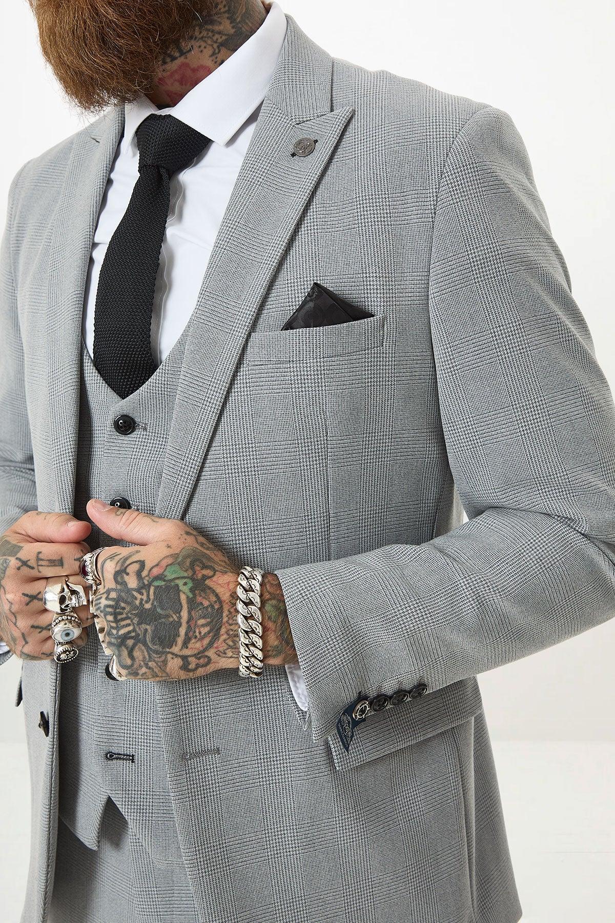 A man wearing the Marc Darcy BROMLEY Silver Grey Check Blazer with a black tie and white shirt adjusts his jacket, revealing tattooed hands decorated with rings and bracelets. Only his torso and lower face are visible.