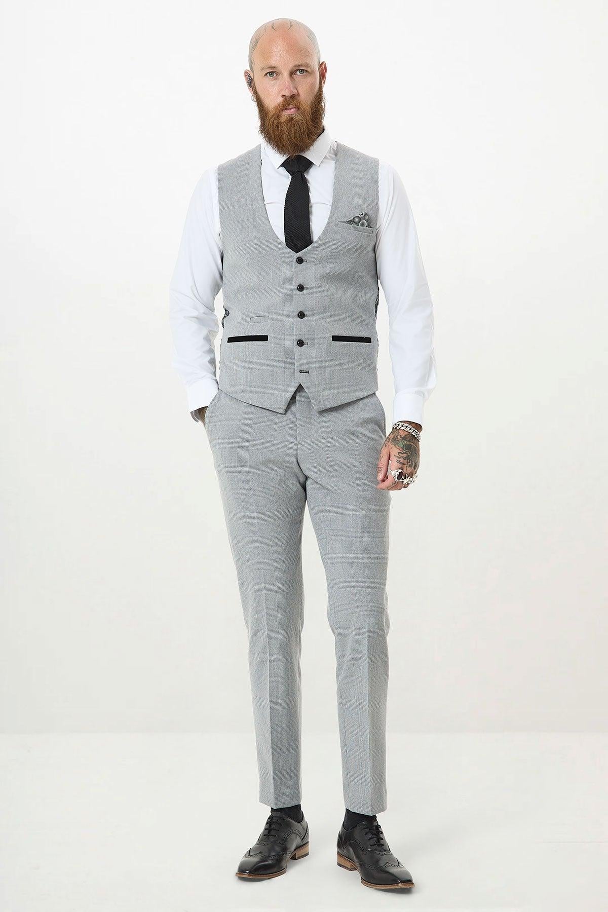 A bearded man with tattoos stands facing forward, hands in pockets, wearing the Marc Darcy BROMLEY Silver Grey Single Breasted Check Waistcoat over a white shirt and black tie, paired with black dress shoes. The background is plain white.