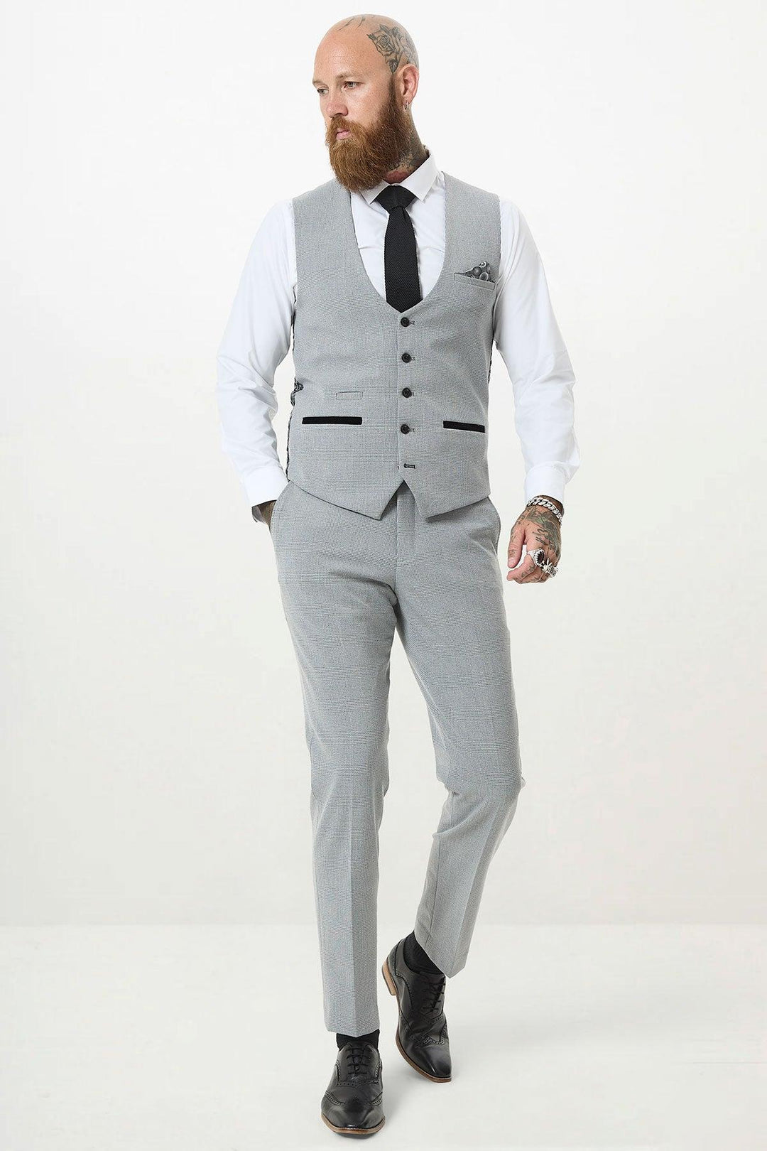A bearded man with tattoos walks confidently against a white background, wearing the Marc Darcy BROMLEY Silver Grey Single Breasted Check Waistcoat in a slim fit, paired with a white shirt, black tie, and black shoes.