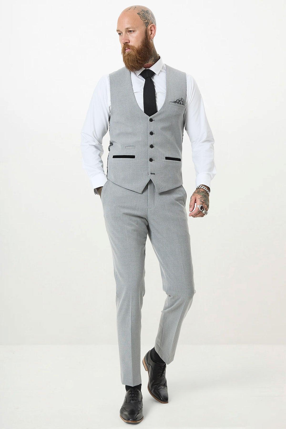 A bearded man in a Marc Darcy BROMLEY silver grey single-breasted check waistcoat, light gray suit, white shirt, black tie, and black shoes stands against a plain white background, gazing to the side with one hand in his pocket.