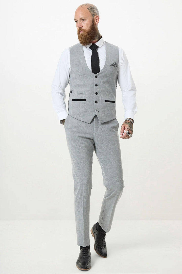 A bearded man with tattoos walks confidently against a white background, wearing the Marc Darcy BROMLEY Silver Grey Single Breasted Check Waistcoat in a slim fit, paired with a white shirt, black tie, and black shoes.