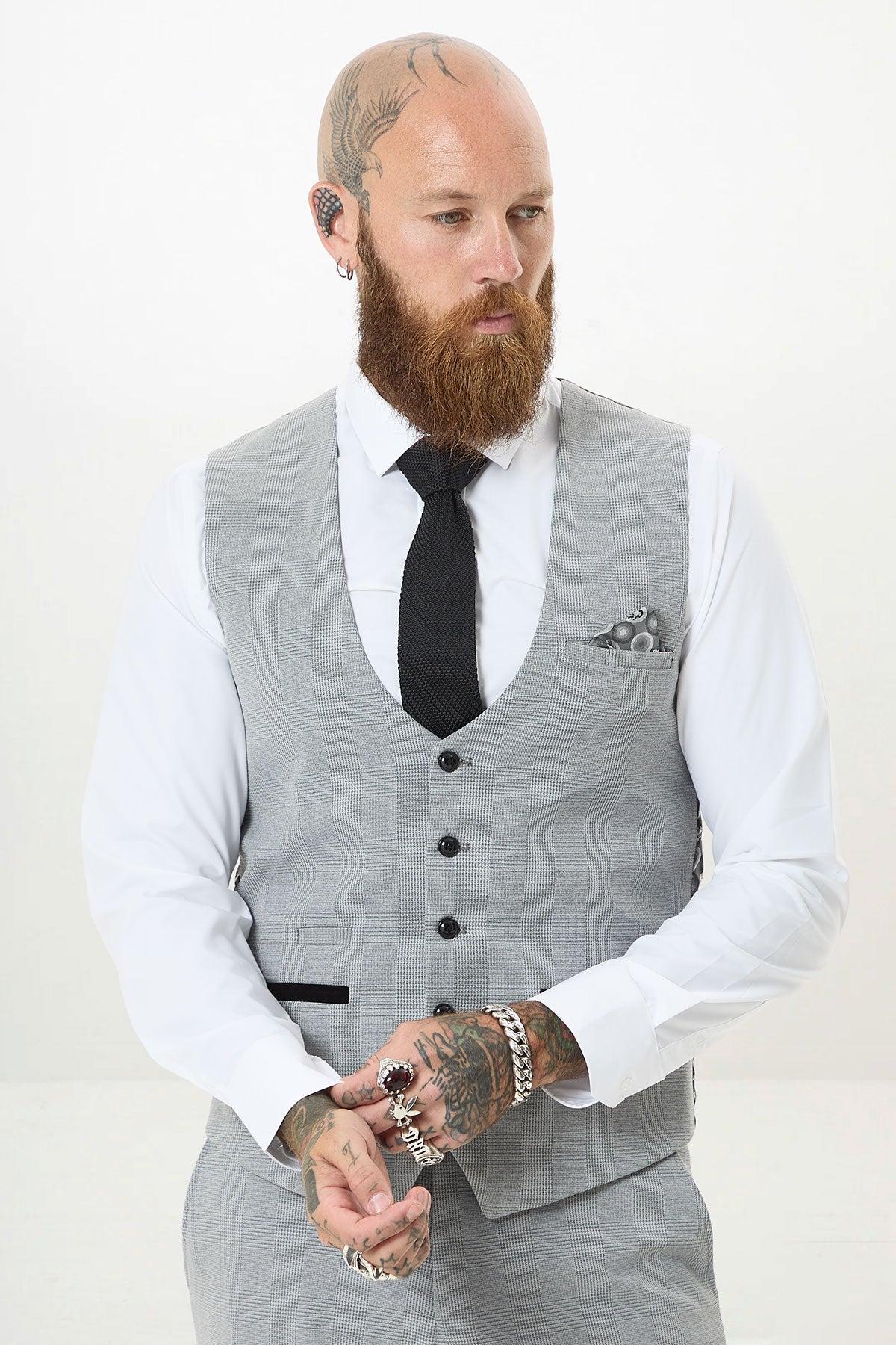A bearded man with tattoos and piercings wears the Marc Darcy BROMLEY Silver Grey Single Breasted Check Waistcoat, paired with matching pants, a white shirt, and black tie. He stands against a plain white background, looking to the side with hands clasped.