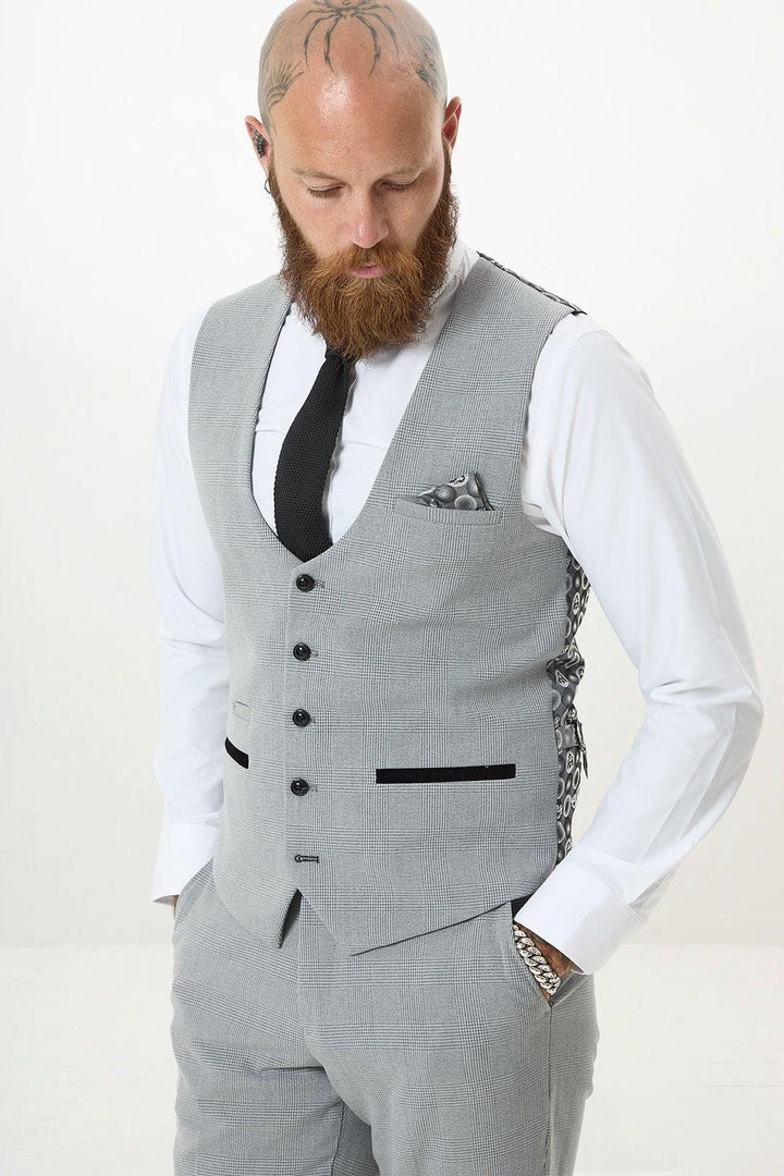 A bald man with a beard wears the Marc Darcy BROMLEY Silver Grey Single Breasted Check Waistcoat, styled slim-fit with matching suit pants, white shirt, black tie, and pocket square; he looks down with hands in his pockets against a plain white background.