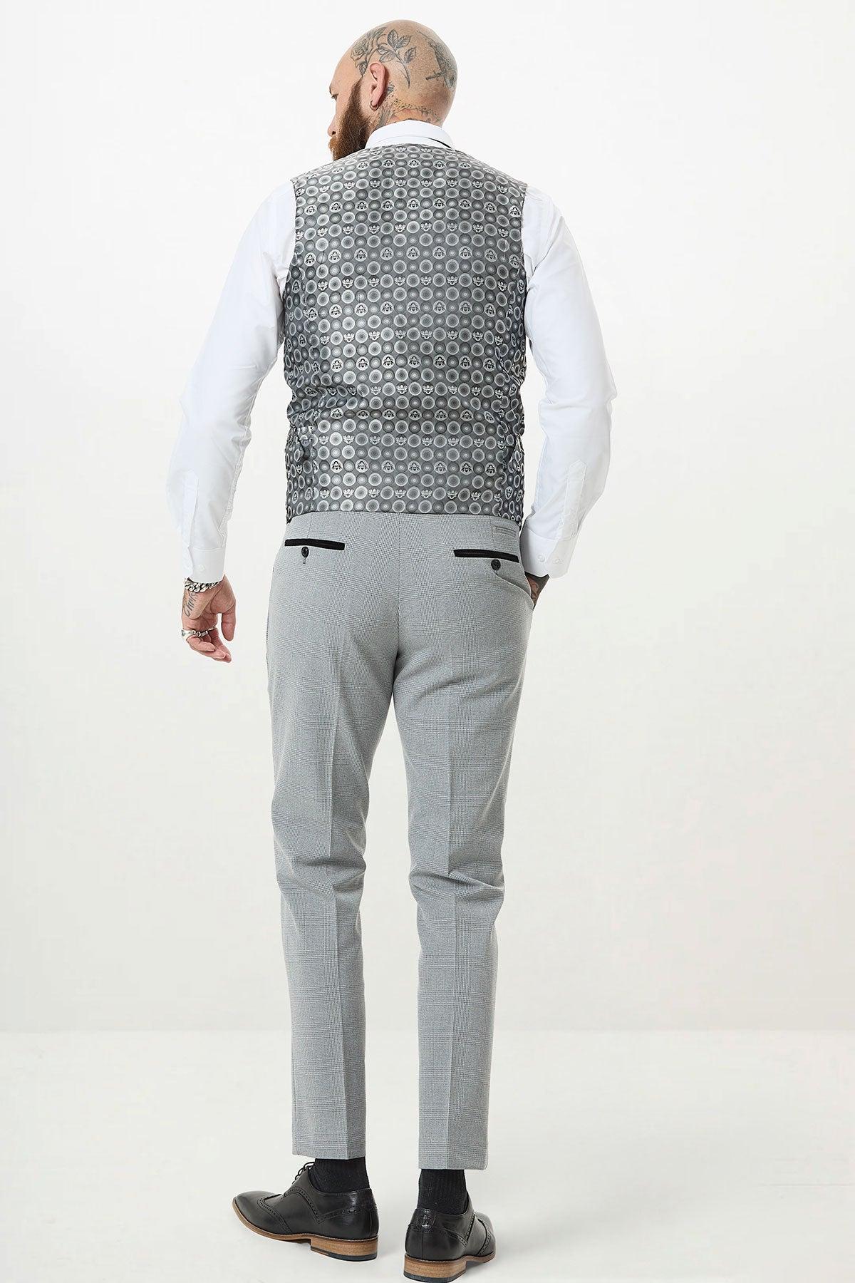 A man with head and hand tattoos stands facing away, wearing a white shirt, Marc Darcy's BROMLEY Silver Grey Single Breasted Check Waistcoat, light grey trousers, and black dress shoes against a plain white background.