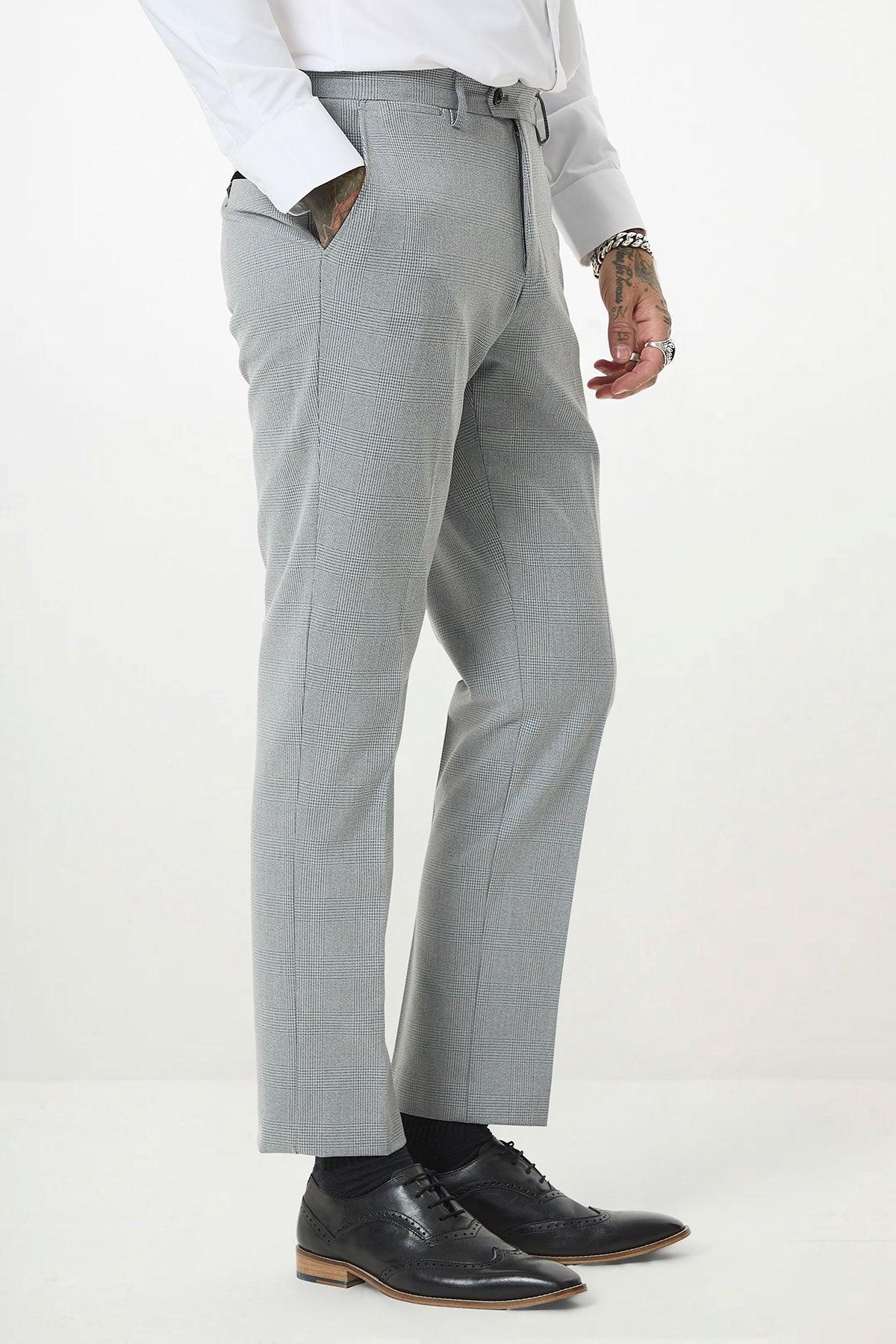 A person wears Marc Darcy’s BROMLEY - Silver Grey Check Trousers with a white shirt, black socks, and black dress shoes, standing with one hand in their pocket and showcasing tattoos on their left hand and wrist.