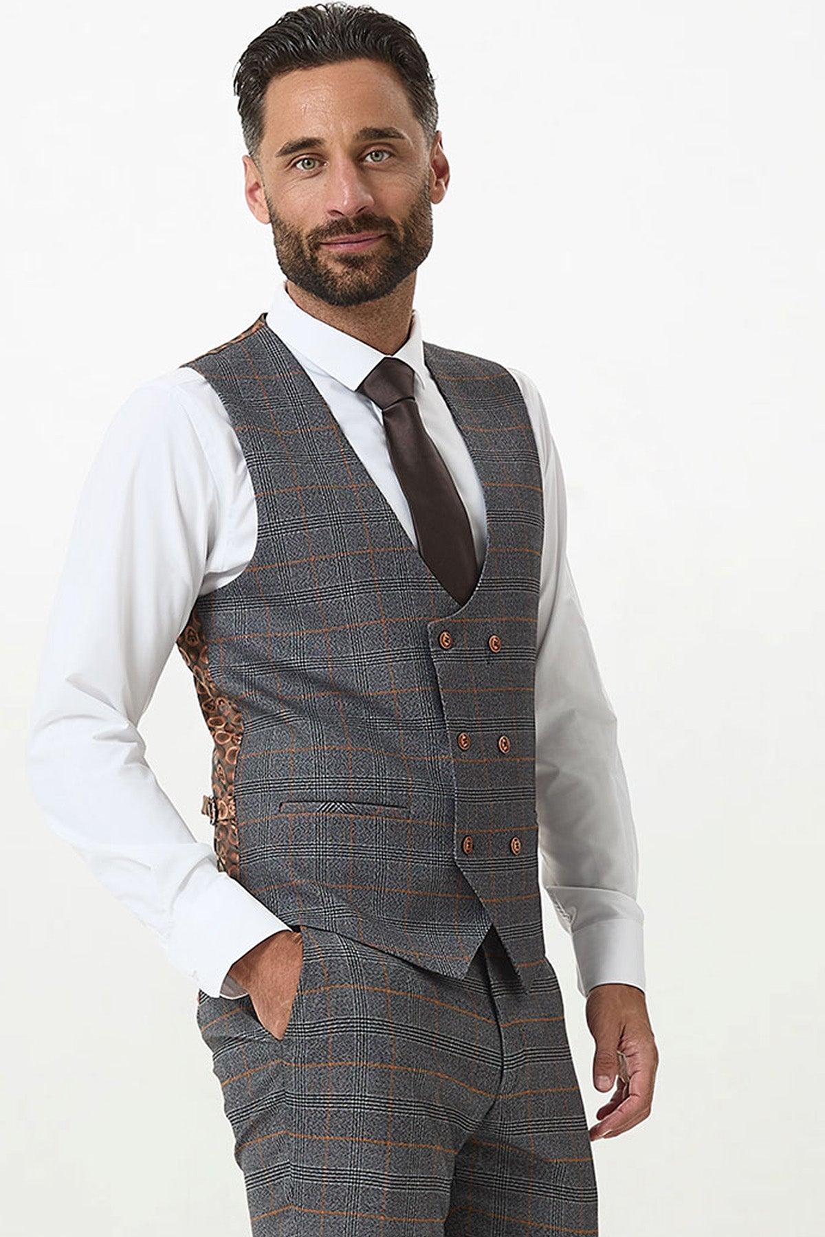 A dark-haired, bearded man wears a white shirt, brown tie, and the marcdarcy JENSON Grey Check Double Breasted Waistcoat with matching trousers, posing with one hand in his pocket against a plain light background.
