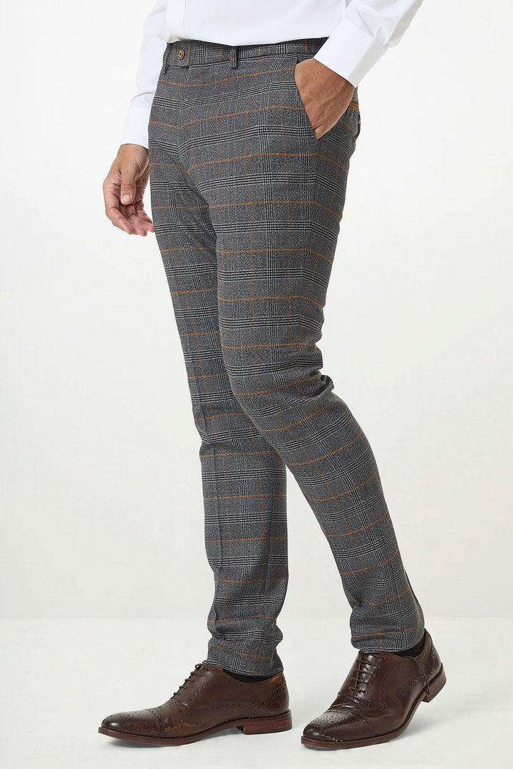A man wearing marcdarcys JENSON - Grey Check Trousers with an orange check pattern, styled with a white shirt and brown brogue dress shoes, stands against a plain white background.