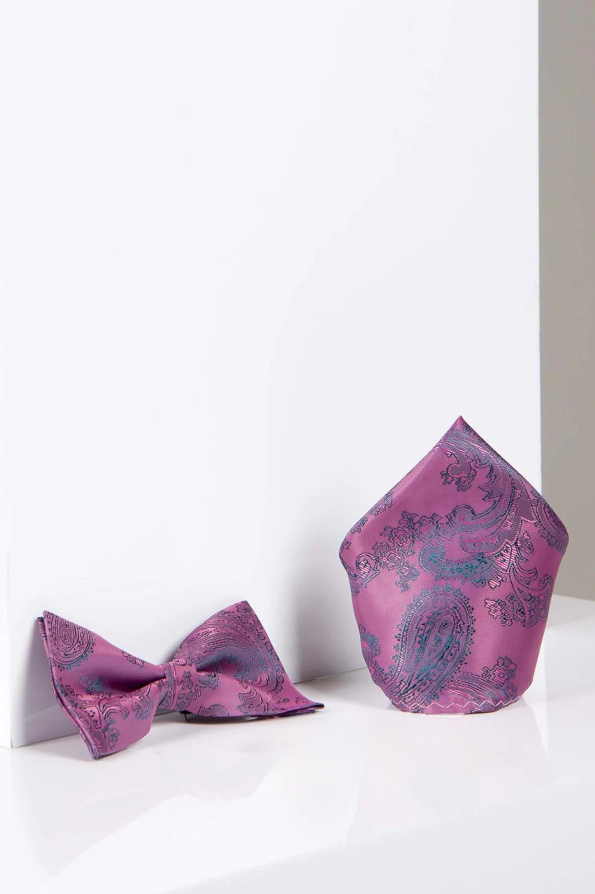 The Accessory PAISLEY Pink Paisley Bow Tie and Pocket Square Set features a folded pink paisley pocket square standing beside a matching bow tie on a glossy white surface, ideal for an elegant evening look.