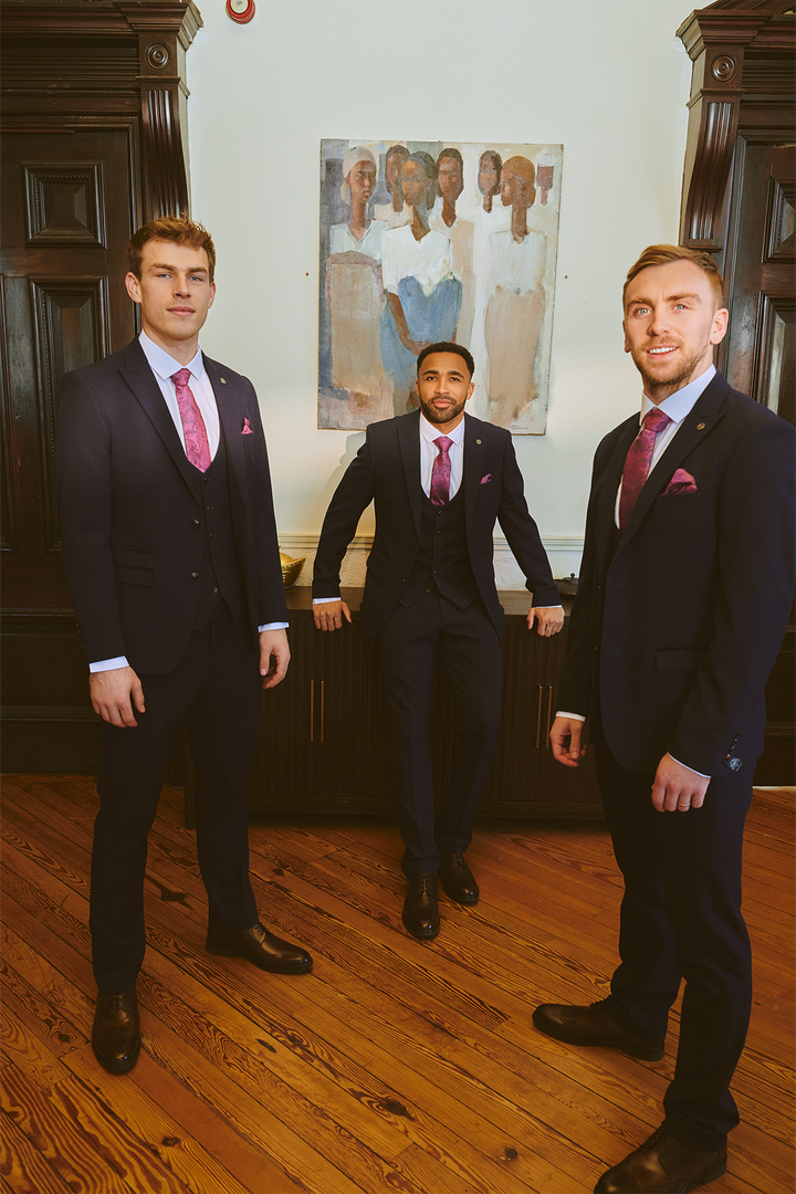 Three men in dark suits, including the Marc Darcy West Ham United Club Suit | BROMLEY Navy Three Piece Suit, pose in a formal room with wooden floors and dark wood paneling; one leans on a cabinet while others stand beneath a painting.