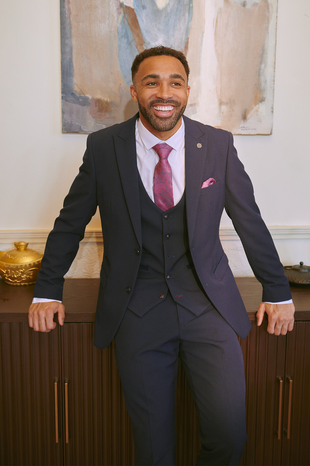 A man in a Marc Darcy BROMLEY Navy Three Piece Suit, as worn by Callum Wilson, smiles while leaning against a brown cabinet topped with elegant decor, with abstract art displayed on the wall behind him.