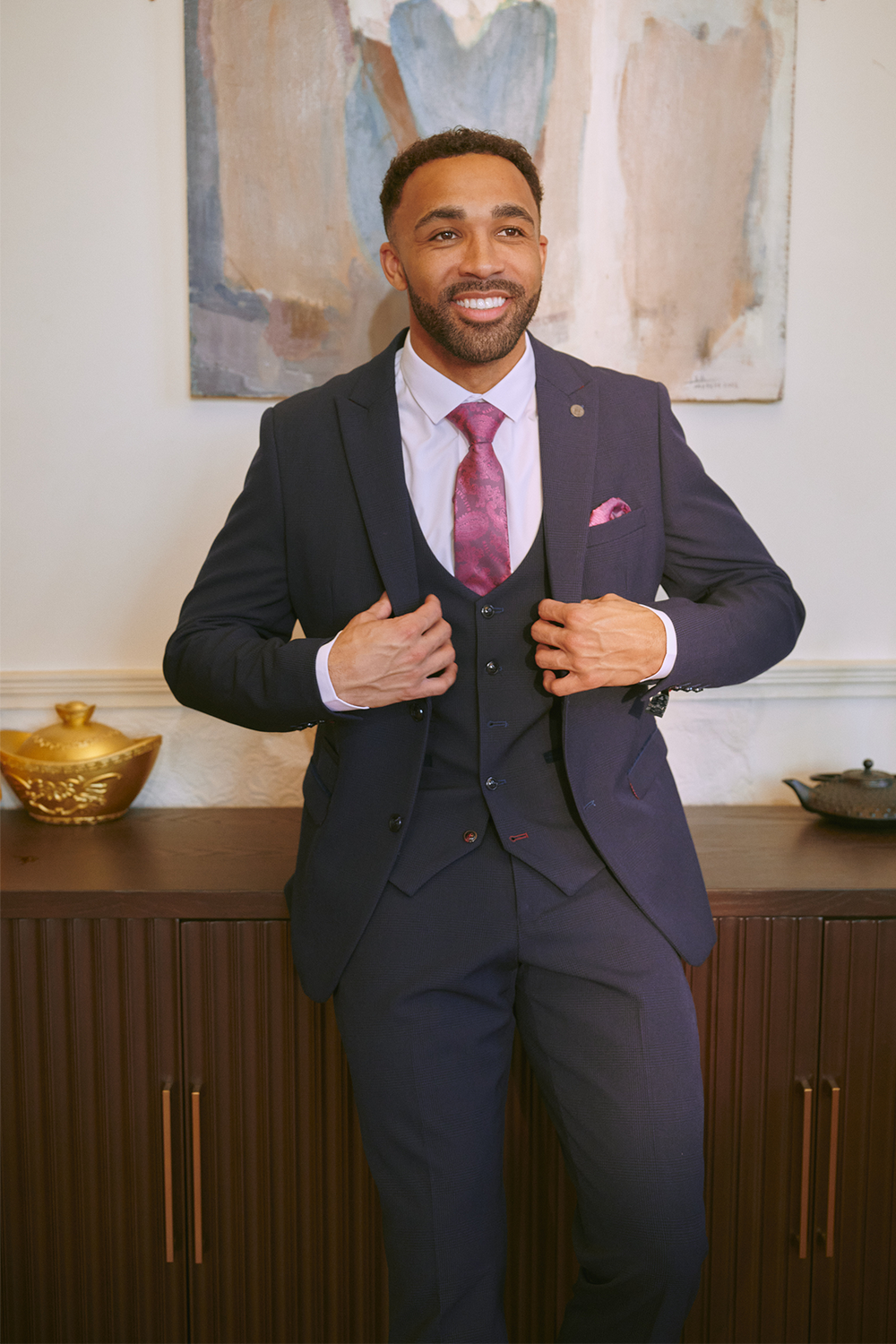 Callum Wilson smiles in the Marc Darcy BROMLEY Navy Three Piece Suit, styled with a pink tie and pocket square, standing before a wooden sideboard adorned with decorative bowls and abstract artwork.