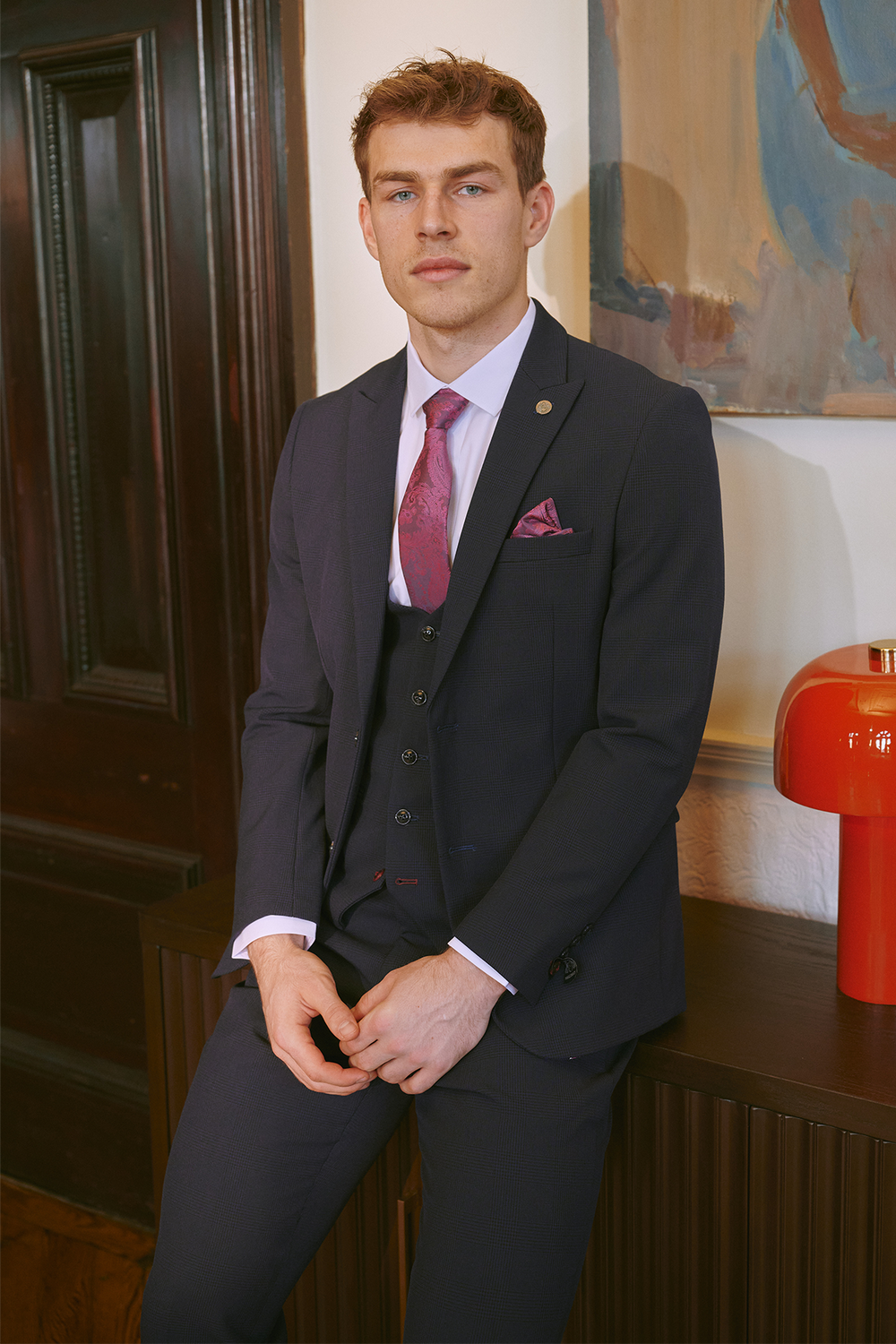 A man in the Marc Darcy BROMLEY Navy Three Piece Suit, as worn by Mads Hermansen, pairs it with a pink shirt, patterned tie, and pocket square while sitting on a cabinet beside a red lamp and dark wooden door.