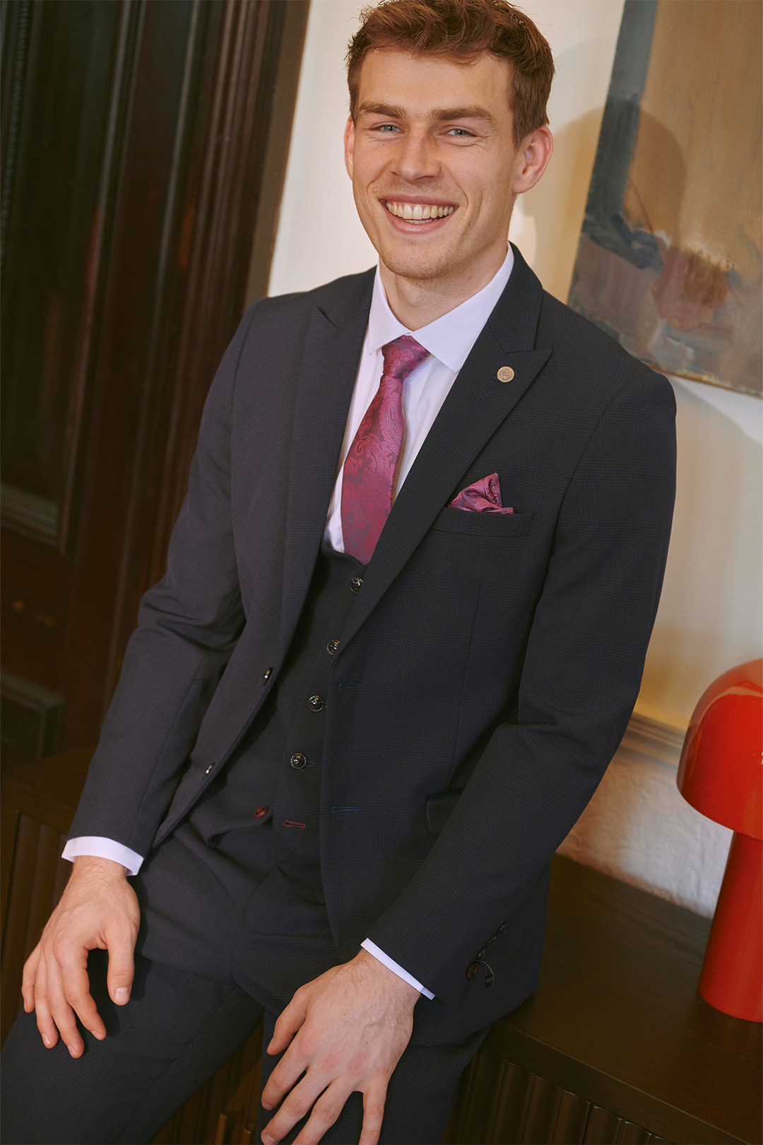 A man with short brown hair wears the Marc Darcy BROMLEY Navy Three Piece Suit, as worn by Mads Hermansen, styled with a pink tie and pocket square. He sits indoors on a table, with a red lamp and abstract painting in the background.