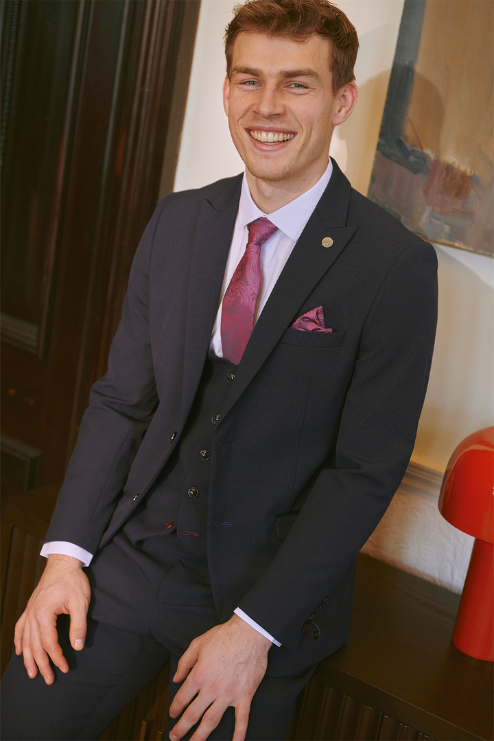 A young man in the Marc Darcy BROMLEY Navy Three Piece Suit, white shirt, and pink tie with matching pocket square sits on a bench and smiles at the camera. A red lamp and an abstract painting are visible in the background.