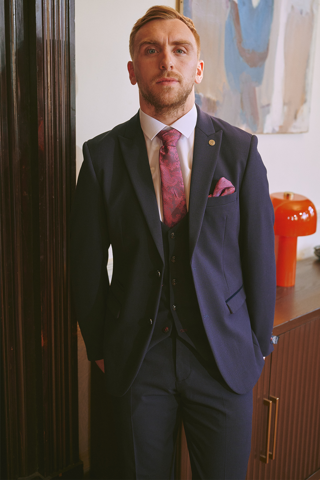 A man with short blond hair, beard, and mustache stands indoors wearing the Marc Darcy BROMLEY Navy Three Piece Suit—As Worn By Jarrod Bowen—with a white shirt and pink tie, in front of a brown cabinet, modern art, and a red lamp.