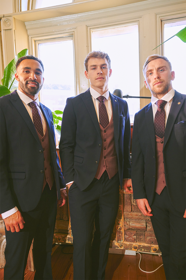 Three men pose indoors in Marc Darcy’s BROMLEY Navy With Monty Waistcoat Suit—as worn by Mads Hermansen—paired with white shirts and patterned ties. Green plants and exposed brick are seen behind them.