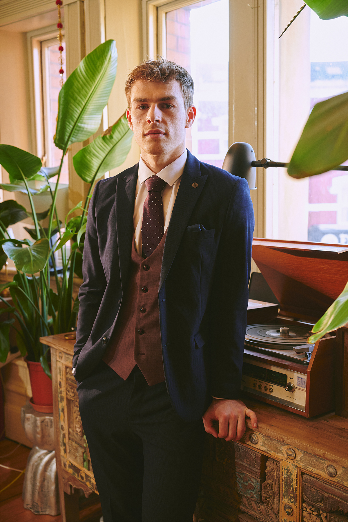 A young man in a Marc Darcy BROMLEY Navy With Monty Waistcoat Suit, as worn by Mads Hermansen, stands indoors by a vintage record player, surrounded by green potted plants and sunlight from large windows.