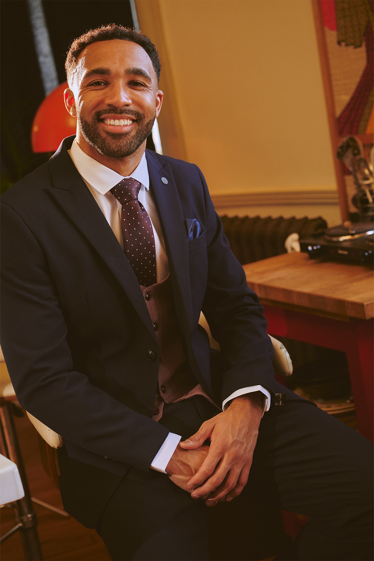 Callum Wilson wears the Marc Darcy BROMLEY Navy With Monty Waistcoat Suit, polka dot tie, and vest. Seated indoors on a chair, he smiles at the camera with short curly hair and a trimmed beard. A wooden table and colorful artwork appear behind him.