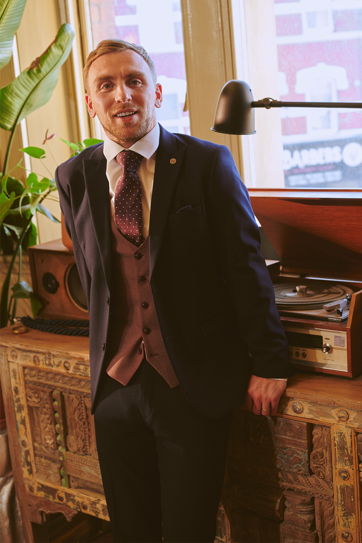 A man smiles indoors, leaning on an ornate cabinet with a vintage record player and green plant, wearing the Marc Darcy BROMLEY Navy With Monty Waistcoat Suit as worn by Jarrod Bowen. Warm sunlight streams through the window.