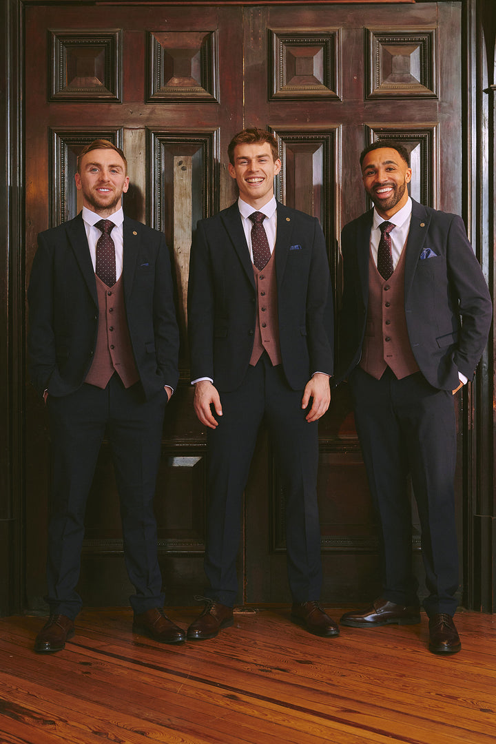 Three men in matching Marc Darcy BROMLEY Navy With Monty Waistcoat Suits, as worn by Mads Hermansen, and patterned ties smile together in front of a large dark wooden door on a polished wood floor.