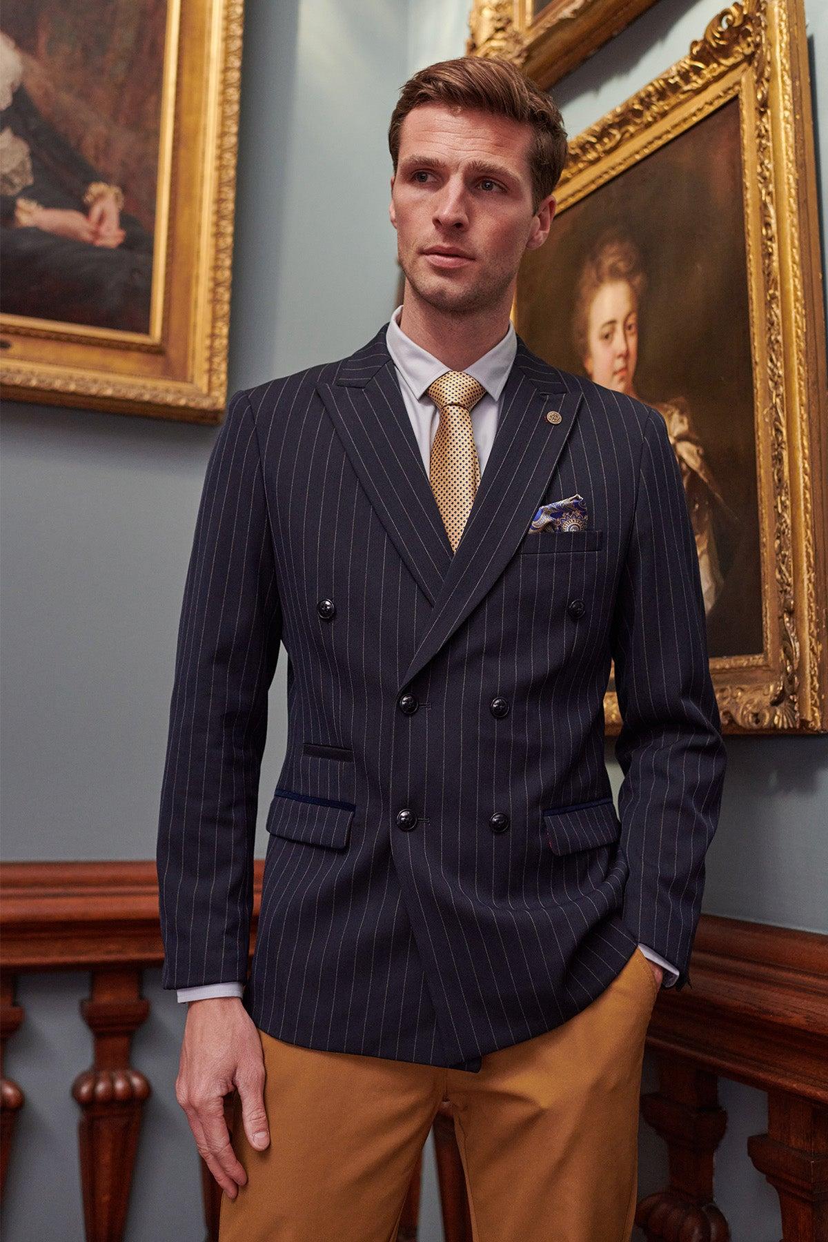 A man in the Marc Darcy ROCCO Navy Pinstripe Double Breasted Blazer, light blue shirt, and yellow tie stands indoors by a wooden railing, with classic framed portraits and blue walls in the background.