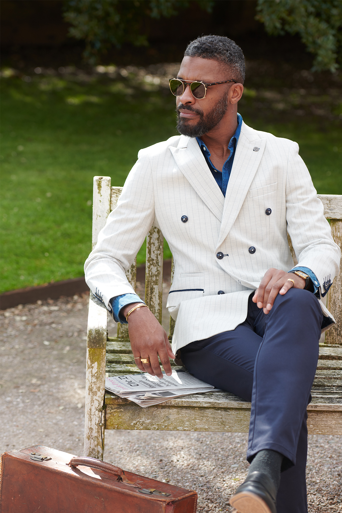 A Black man wears the GRANT Stone Double Breasted Pinstripe Blazer by Marc Darcy with navy pants while sitting on a wooden bench outdoors reading a newspaper.
