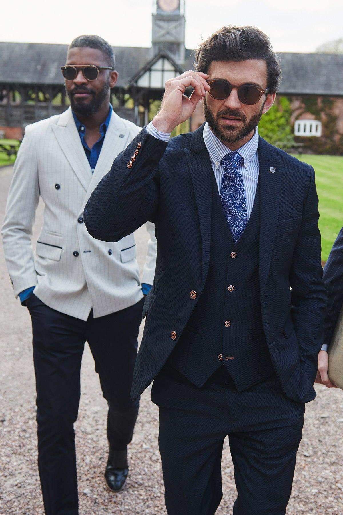 Two men walk outside on a gravel path. The man in front wears the marcdarcy MAX Navy Three Piece Suit with Contrast Buttons, paired with a patterned tie, adjusting his sunglasses. The man behind wears a light blazer and dark pants; a building and lawn are visible nearby.