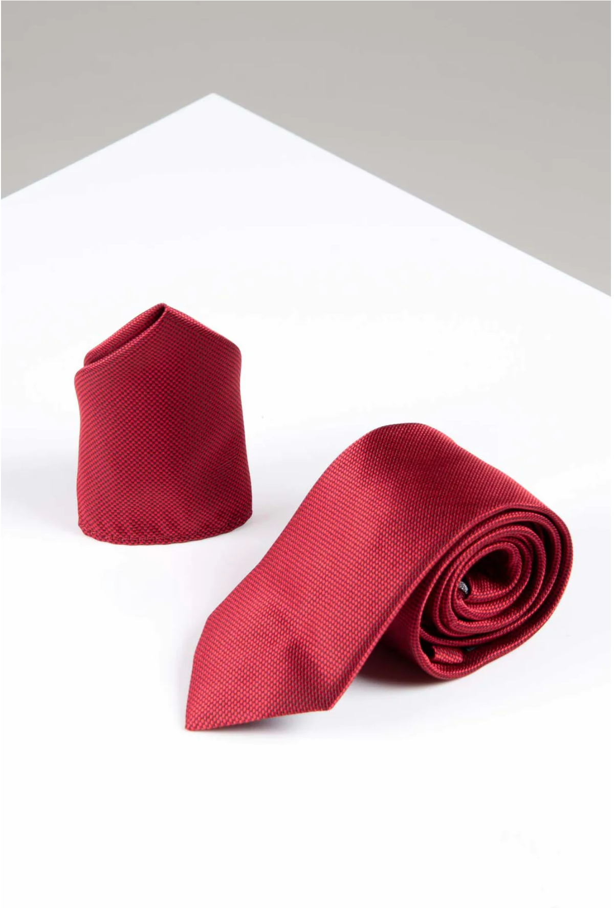 The Accessory TOBY Birdseye Tie and Pocket Square Set in Wine features one tie rolled and another upright, displayed on a white and light gray geometric surface.