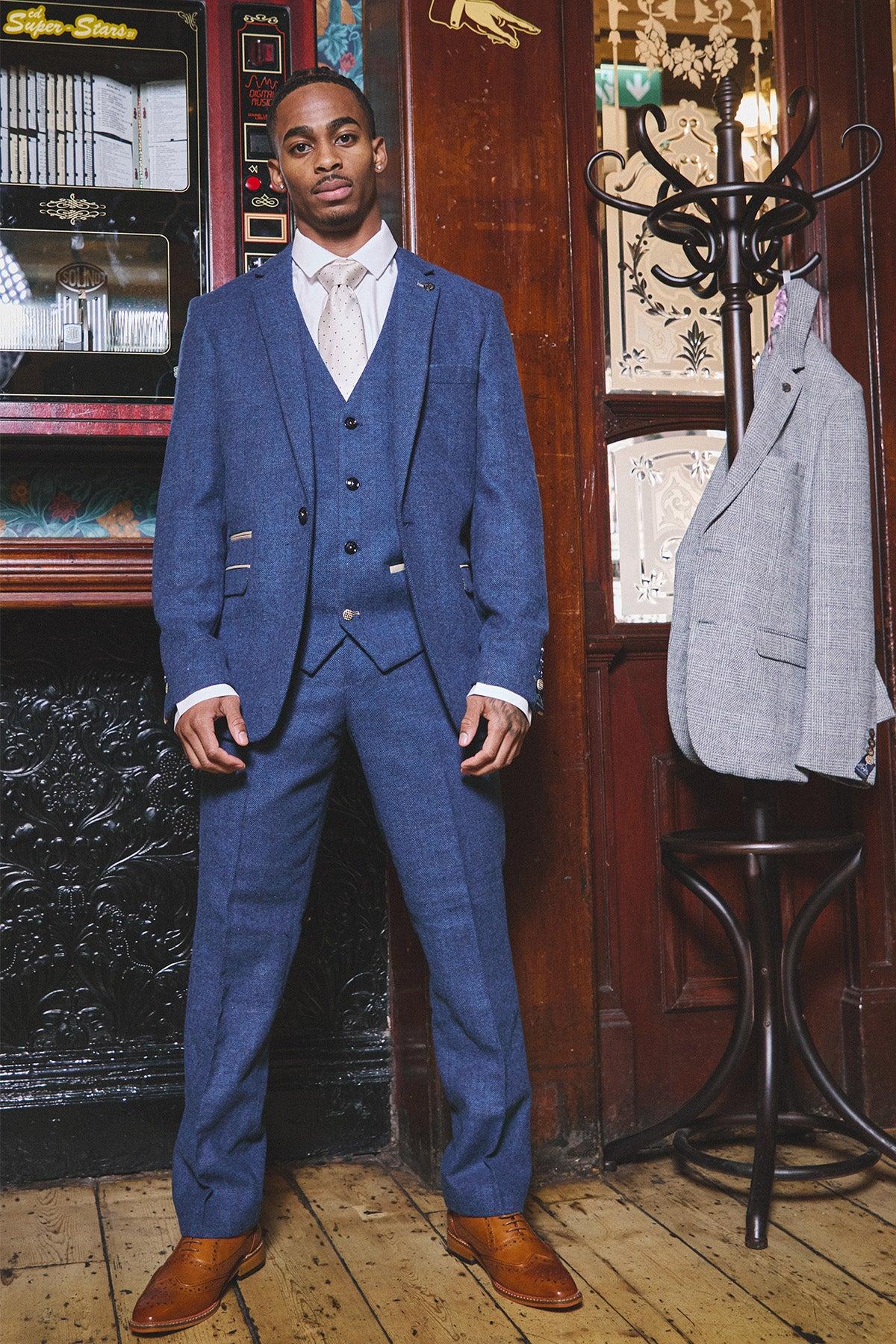 A Black man wears the marcdarcy WHU Collection DION Blue Tweed Check Suit, as worn by Crysencio Summerville, with tan dress shoes, standing next to a coat rack holding a grey tweed jacket.