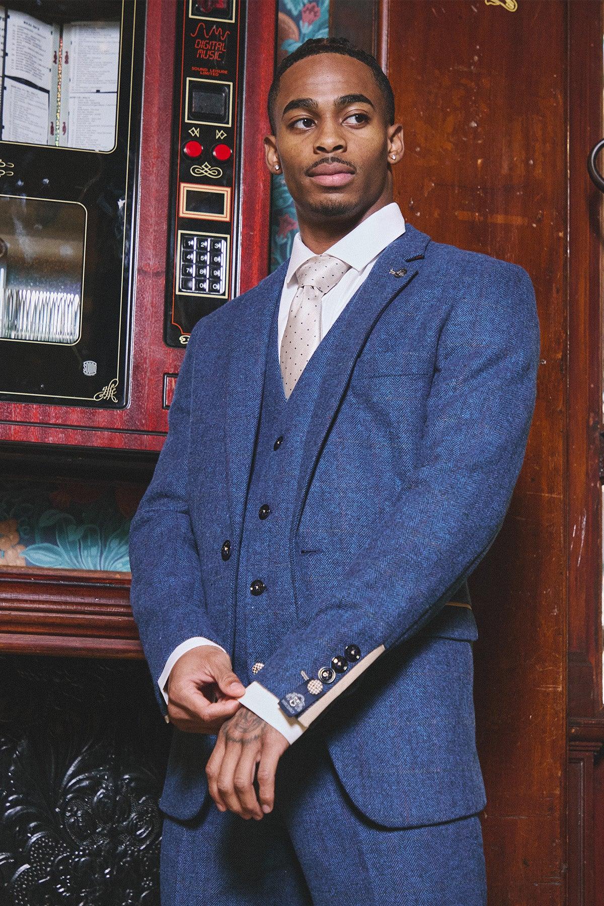 A young Black man wears the marcdarcy WHU Collection - DION Blue Tweed Check Suit as worn by Crysencio Summerville, paired with a white shirt and cream tie, standing before a vintage jukebox.