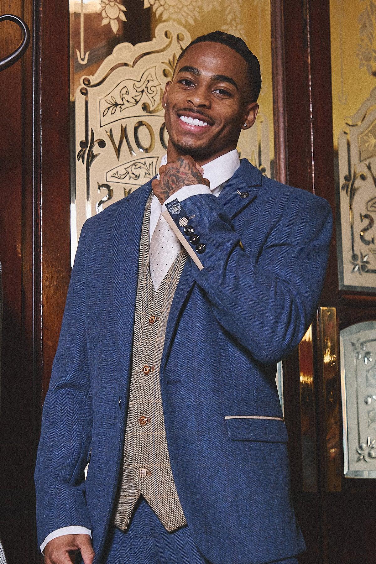 A man smiles and adjusts his collar in front of ornate glass doors, wearing marcdarcy’s WHU Collection – DION Blue Tweed Suit with TED Waistcoat, as worn by Crysencio Summerville.