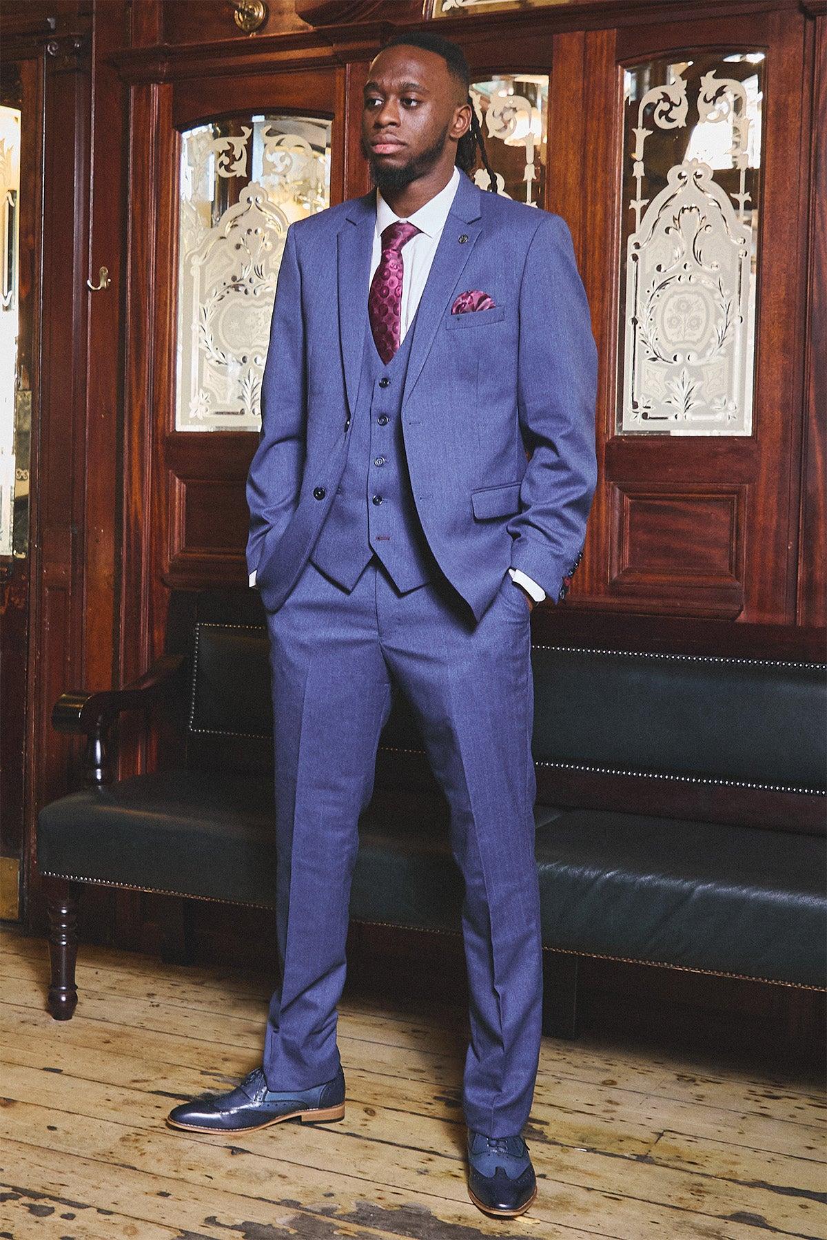 A man in the Marc Darcy WHU Collection - ASHLEY Navy Blue Three Piece Suit, as worn by Aaron Wan-Bissaka, with a maroon patterned tie and pocket square stands on a wooden floor before ornate panels with decorative glass.