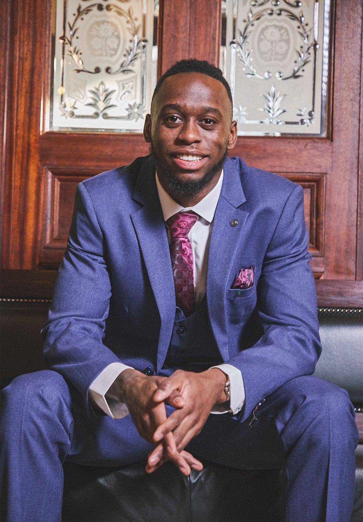 A Black man in a slim fit Marc Darcy WHU Collection ASHLEY Navy Blue Three Piece Suit, white shirt, and patterned tie smiles while sitting in front of a wooden door with decorative glass panels.