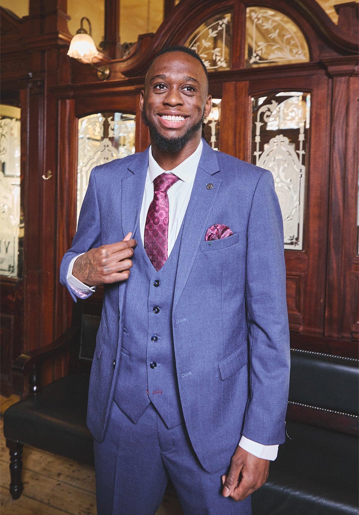 A Black man in the Marc Darcy WHU Collection - ASHLEY Navy Blue Three Piece Suit, as worn by Aaron Wan-Bissaka, with a white shirt and burgundy tie, smiles broadly before wooden panels and ornate glass doors, embodying timeless formalwear style.