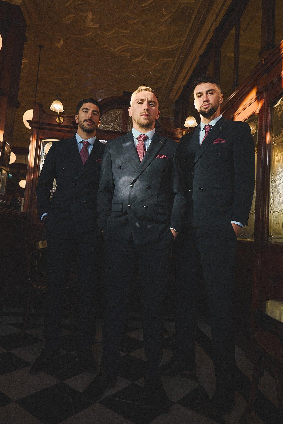 Three men in dark suits and ties, including the Marc Darcy WHU Collection - BROMLEY Navy Double Breasted Suit as worn by Jarrod Bowen, stand confidently in an elegant vintage-inspired room with ornate ceilings, mirrors, and ambient lighting.